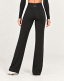 A model from the back wearing black high-waisted wide-leg pants with a small silver 'HR' logo on the waistband, paired with a black long-sleeve top and white shoes, against a light gray studio background.