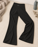 Black wide-leg pants laid flat on a beige fabric surface, with a knit blanket, dried leaves, and HER Stone Water bottle as styled props. 