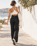 A woman walking away on a sunny seaside path wearing black high-waisted athletic pants and a white sports bra, carrying a cream canvas tote bag.